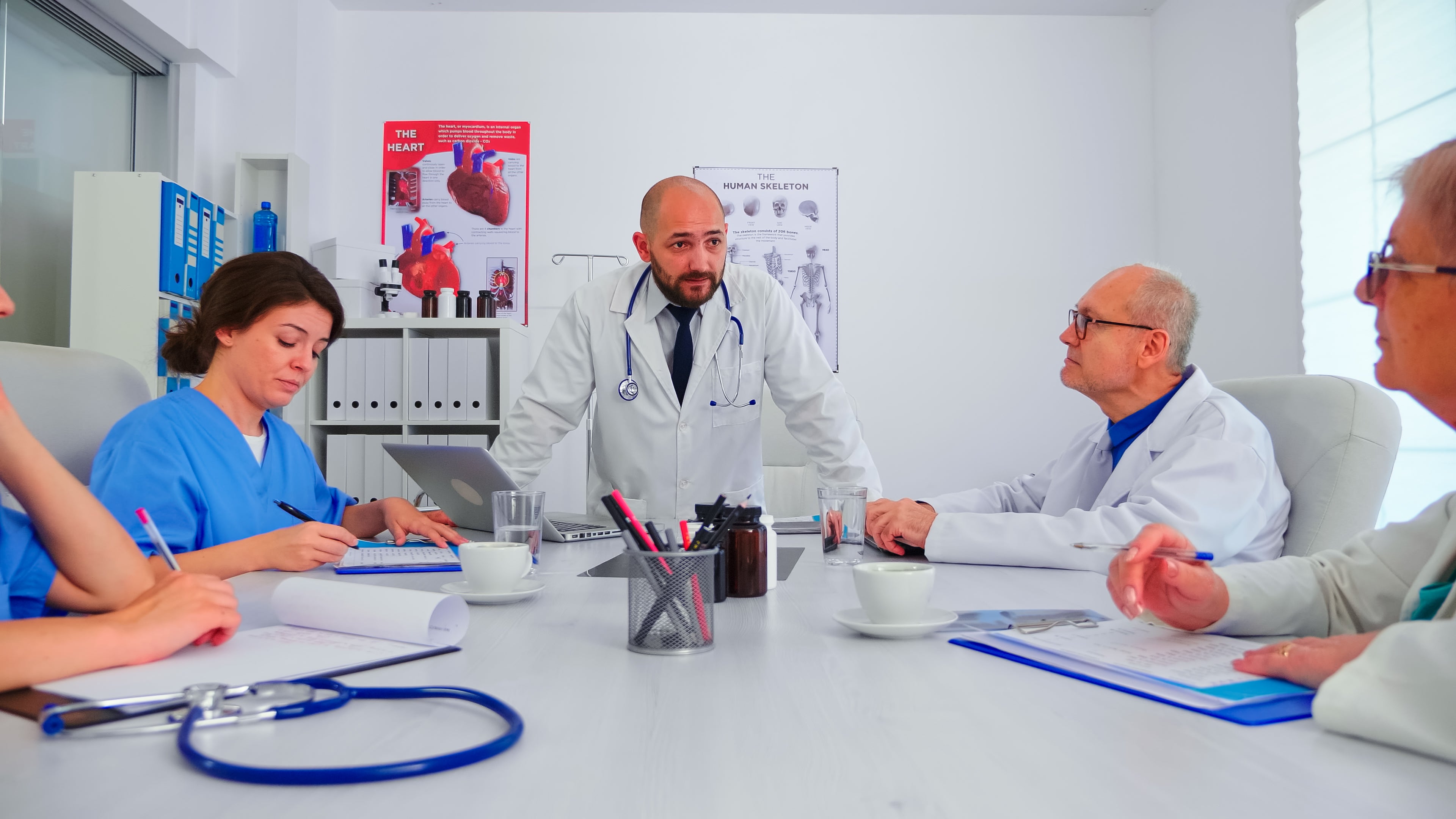 A group of medical professionals, including doctors in white coats and a nurse, have a discussion around a table in a clinical meeting room.